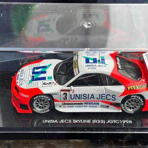 Könnte beinhalten: Ein 1:43 Druckguss-Modell eines weiß-roten Unisia Jecs Nissan Skyline R33 Rennwagens aus der JGTC-Saison 1998. Das Modell befindet sich in einer klaren Acrylvitrine mit schwarzem Sockel und trägt die Aufschrift Unisia Jecs Skyline R33 JGTC 1998.
