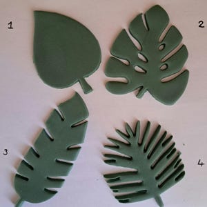 May include: Four green leaf-shaped decorations. The leaves vary in shape, including a solid oval, a split leaf, and two different types of fern-like leaves. The leaves are all a solid, muted green colour and appear to be made of a pliable material.