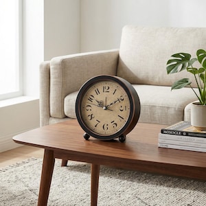 May include: A round, vintage-style clock with a dark brown frame and a cream-colored face. The clock features black numerals and hands. It is displayed on a wooden coffee table, alongside a potted plant and books. The clock is approximately 8 inches in diameter.