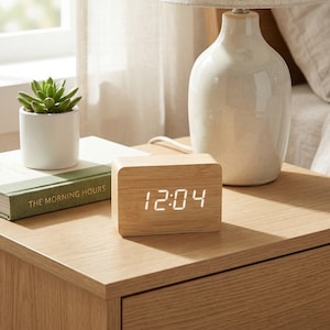 May include: A wooden digital clock displaying 12:04 sits on a wooden nightstand. A white ceramic lamp and a small potted succulent are also on the nightstand. A book titled "The Morning Hours" is visible.