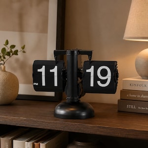 May include: A black retro flip clock showing "11:19 PM". The clock has a round base and a central support with two panels displaying the time. The numbers are white against a black background. The clock is on a wooden surface.