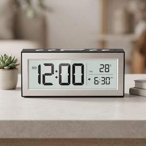 Puede incluir: Un despertador digital con marco plateado y parte superior negra. La pantalla muestra la hora como 12:00 AM, la fecha y la hora de la alarma a las 6:30 AM. La temperatura se muestra a 28 grados Celsius.