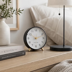 May include: A black and gold analog alarm clock with a white face, gold legs, and gold hands. The clock is on a wooden surface with a lamp and a vase in the background. The clock face has the text "Designed by JBT".