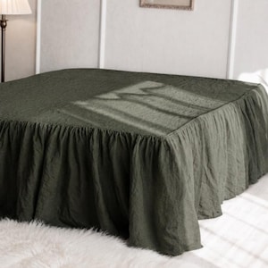 May include: A dark olive green bed skirt with a gathered, ruffled edge that drapes down to the floor. The fabric has a textured appearance and covers the base of the bed, extending from the top surface down to the bottom edge of the frame.