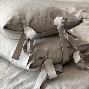 May include: Two rectangular pillows made of light beige linen fabric. Each pillow features a series of fabric ties along one edge, designed to be knotted into bows. The texture of the linen is visible, showing a natural weave and soft appearance.