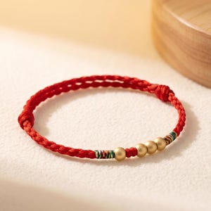 May include: Red braided bracelet with gold-colored beads and colorful accents. The bracelet is made of a woven red cord and features three gold beads and decorative bands with green, blue, and yellow threads.
