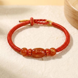 Pode incluir: Pulseira vermelha trançada com três contas vermelho-alaranjadas. A conta central, oval, tem símbolos dourados. A pulseira é atada com detalhes dourados.
