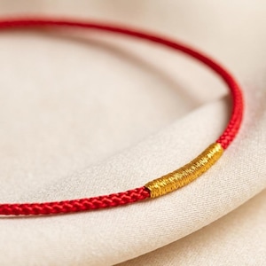 May include: A close-up of a red cord necklace with a gold-colored cylindrical clasp. The necklace is set against a soft, off-white fabric background. The cord appears to be made of woven threads.