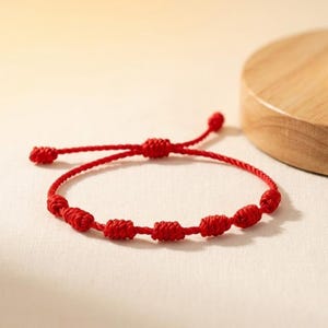 Può includere: Un braccialetto di corda rossa con più nodi. Il braccialetto è regolabile e presenta una serie di nodi distanziati uniformemente lungo la sua lunghezza. Lo sfondo è di un colore morbido e neutro, che mette in risalto il rosso vibrante del braccialetto.