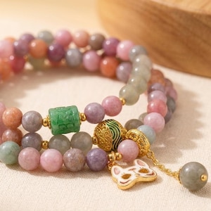 Puede incluir: Una pulsera de múltiples hilos con cuentas en tonos rosa, morado y gris. La pulsera incluye un cilindro de jade verde, detalles dorados y un dije con un diseño de zorro blanco y dorado. Una cuenta gris cuelga de una cadena dorada.