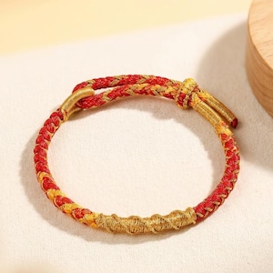 Op de afbeelding: Een gevlochten armband met rode en gouden draden. De armband heeft een geweven ontwerp met een knoopsluiting. De armband heeft een cirkelvorm en wordt waarschijnlijk om de pols gedragen.