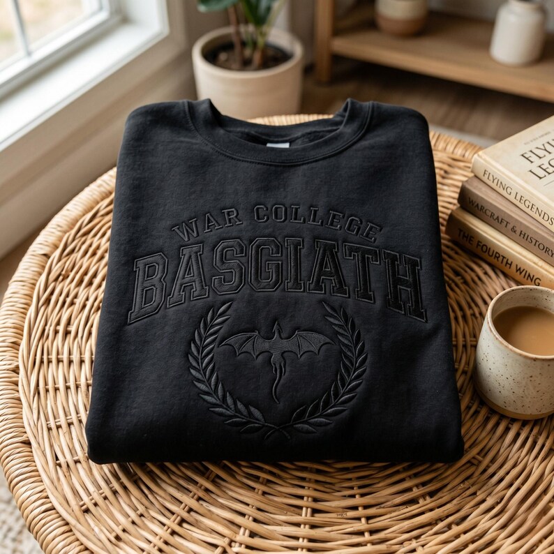 K&ouml;nnte beinhalten: Schwarzes Sweatshirt mit dem Text "WAR COLLEGE BASGIATH" im College-Stil. Darunter befindet sich ein Drachenemblem in einem Lorbeerkranz. Das Sweatshirt liegt gefaltet auf einem Korbtisch.