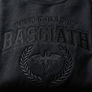 K&ouml;nnte beinhalten: Schwarzes Sweatshirt mit dem Schriftzug "WAR COLLEGE BASGIATH" in einer College-Schriftart. Darunter befindet sich ein Drachenemblem in einem Lorbeerkranz. Das Design ist Ton-in-Ton gestickt.