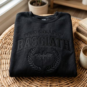 K&ouml;nnte beinhalten: Schwarzes Sweatshirt mit Rundhalsausschnitt und dem Text "WAR COLLEGE BASGIATH" im College-Stil. Darunter befindet sich ein Drachenemblem in einem Lorbeerkranz. Das Sweatshirt liegt auf einer Korboberfl&auml;che.