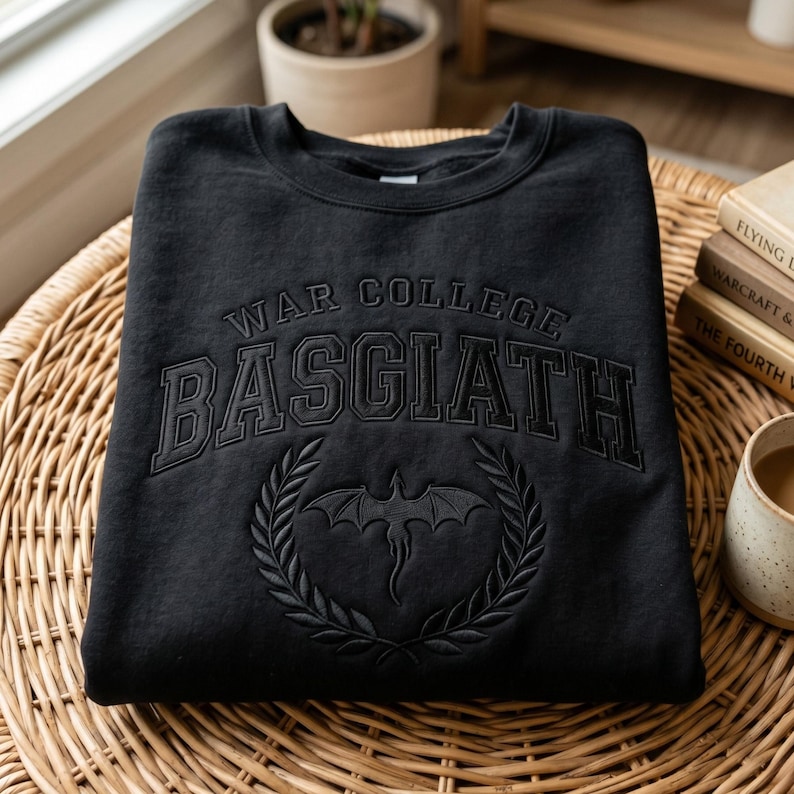 K&ouml;nnte beinhalten: Schwarzes Sweatshirt mit Rundhalsausschnitt und dem Text "WAR COLLEGE BASGIATH" im College-Stil. Darunter befindet sich ein Drachenemblem in einem Lorbeerkranz. Das Sweatshirt liegt auf einer Korboberfl&auml;che.