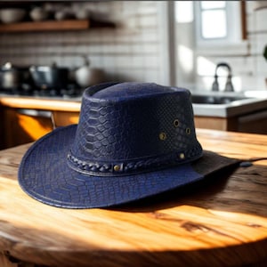 May include: A navy blue cowboy hat with a textured, snakeskin-like pattern. The hat features a braided band and small gold-colored rivets around the crown. The brim is wide and the hat sits on a wooden table.