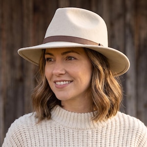 May include: A cream-colored fedora hat featuring a structured crown and a wide brim. The hat is accented with a thin, dark brown leather band encircling the base of the crown, providing a distinct contrast to the light-colored felt material.