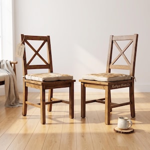 May include: Two wooden chairs with a medium brown finish, featuring an X-shaped backrest design and light-colored seat cushions. A small tag is attached to the left chair, and the text Rustique House Co. is visible on the right chair frame.