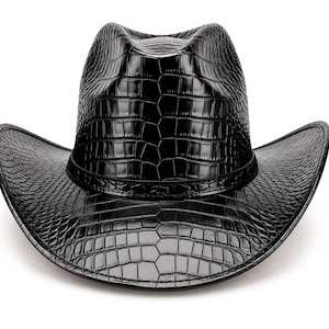 May include: A black cowboy hat featuring a glossy, embossed crocodile skin texture across the entire surface. The hat has a structured crown with a central crease and a wide, curved brim, accented by a matching black band around the base.