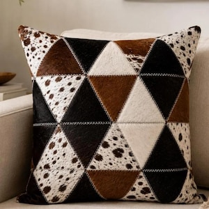 Puede incluir: Un cojín cuadrado con un diseño geométrico de patchwork compuesto por paneles triangulares. El patrón alterna entre negro sólido, marrón sólido y blanco con texturas moteadas estilo piel de vaca y costuras visibles.