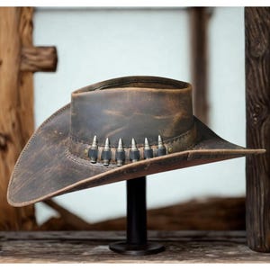 Puede incluir: Sombrero de vaquero de cuero marrón desgastado, con ala ancha y una banda decorativa con seis adornos plateados en forma de bala. El sombrero se exhibe sobre un soporte negro, mostrando su forma y textura. El cuero tiene un aspecto usado.