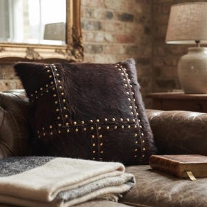 May include: A square dark brown hair-on-hide throw pillow featuring a grid pattern of small metallic studs. The pillow is placed on a tufted brown leather sofa alongside a folded cream and gray woven blanket and a small brown book.