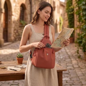 May include: A terracotta-colored sling backpack with a single shoulder strap, featuring a front zippered pocket, a small metallic logo emblem, and matching zipper pulls. The bag is worn across the chest, with a smartphone partially inserted into the top opening.