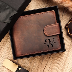 Puede incluir: Cartera de cuero marrón con cierre de botón a presión, presentada en una caja de regalo negra. La cartera está grabada con el nombre "WILLIAM" en una fuente estilizada. El cuero tiene un aspecto envejecido.
