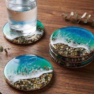 Puede incluir: Un juego de posavasos redondos con un diseño de resina que imita la orilla del mar. Cada posavasos muestra un degradado de tonos azules y verdes que se transforman en olas de espuma blanca, con una base de pequeñas piedras naturales de colores.