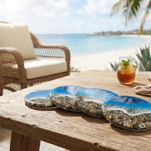 May include: A set of four circular coasters featuring a resin design that mimics a shoreline. Each coaster displays a layer of small, multi-coloured pebbles at the base, transitioning into a clear blue ocean wave with white foam detailing.