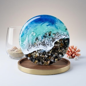 May include: A circular resin art piece featuring a blue and turquoise ocean wave crashing onto a shore of multi-coloured pebbles. The artwork is displayed on a round wooden tray with a gold-toned metal rim, engraved with the text BLOSSOM CAST.