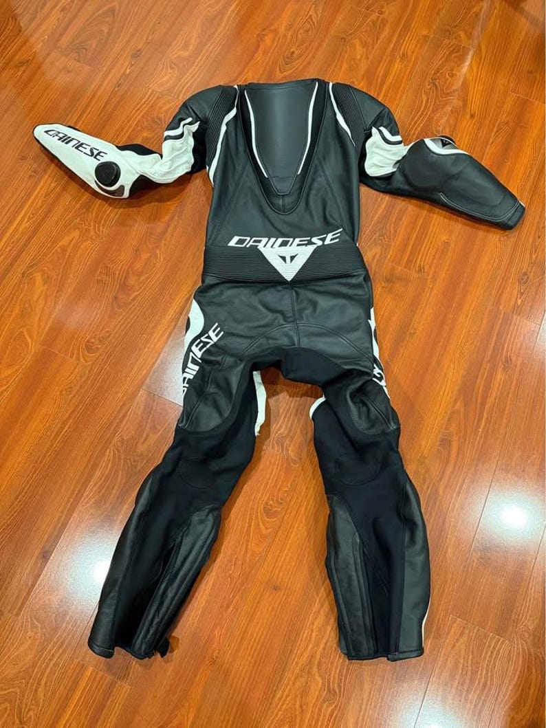 May include: Black and white motorcycle racing suit with the brand name "DAINESE" and "CHINESE" visible. The suit has white accents on the shoulders, arms, and legs. The suit is made of leather and has protective padding.