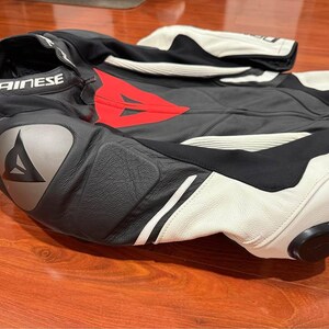 May include: Black and white motorcycle racing suit with red accents. The suit features the "DAINESE" logo on the collar and leg. The suit is made of leather and has protective padding on the shoulders, elbows, and knees.
