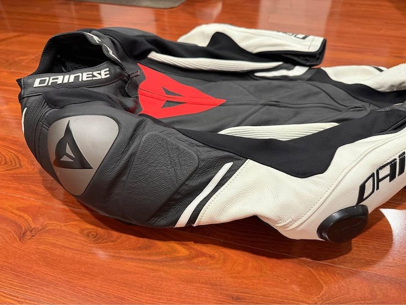 May include: Black and white motorcycle racing suit with red accents. The suit features the "DAINESE" logo on the collar and leg. The suit is made of leather and has protective padding on the shoulders, elbows, and knees.