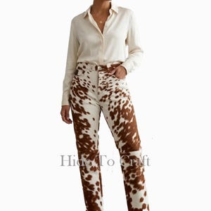 May include: A cream-coloured button-down shirt paired with high-waisted jeans featuring a brown and white cowhide pattern. The jeans have a straight leg cut. The text "Hide To Craft" is visible on the jeans.