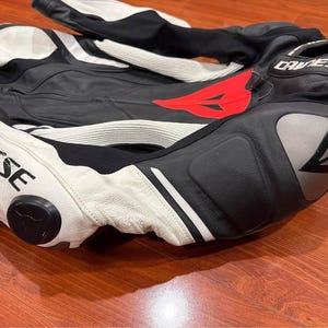 May include: Black and white motorcycle racing suit with red accents. The suit features the brand name "DAINESE" in white lettering. The suit is made of leather and has protective padding on the knees and elbows.