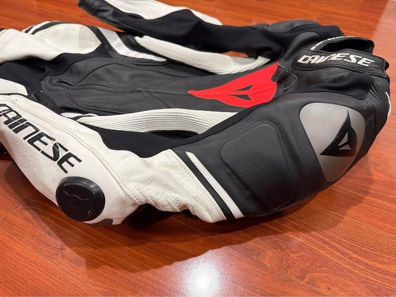 May include: Black and white motorcycle racing suit with red accents. The suit features the brand name "DAINESE" in white lettering. The suit is made of leather and has protective padding on the knees and elbows.