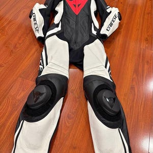 May include: Black and white motorcycle racing suit with red accents. The suit features the brand name "DAINESE" on the sleeves and collar. The suit has protective padding on the knees and elbows.
