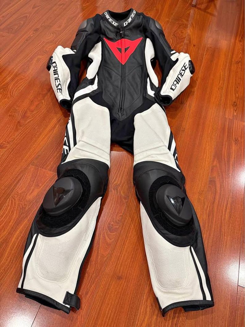May include: Black and white motorcycle racing suit with red accents. The suit features the brand name "DAINESE" on the sleeves and collar. The suit has protective padding on the knees and elbows.