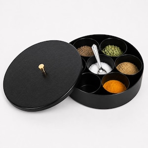 May include: A black spice box with a lid and a gold-coloured knob. The box is round with seven compartments, each filled with different spices. A small spoon rests in one compartment. The lid is open, revealing the spices.