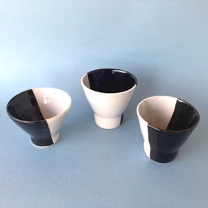 Peut inclure: Trois bols en céramique au design moderne. Chaque bol est blanc avec une bande noire qui s'étend du bord à la base. L'intérieur des bols est blanc ou noir. Les bols sont posés sur un fond bleu clair.