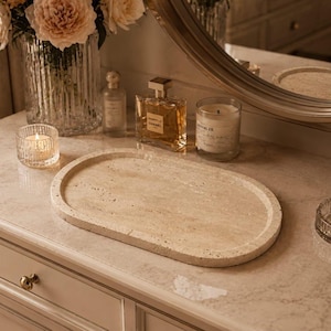 May include: An oval-shaped beige travertine stone tray with a natural porous texture and a raised rim. The tray is positioned on a flat surface, showcasing its smooth, light-coloured surface and distinct organic stone markings throughout the piece.