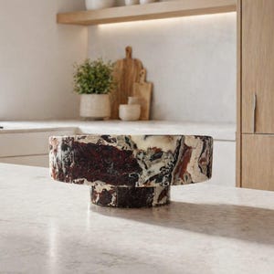 Op de afbeelding: Een decoratieve voetbakschaal met een uniek, gelaagd ontwerp. De schaal heeft een mix van donkerrode, crème- en grijstinten, waardoor een marmerachtig effect ontstaat. Het staat op een lichtgekleurd aanrecht in een keuken.