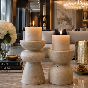 May include: Two cylindrical pillar candles with warm flames resting on top of two stacked, beige, porous stone candle holders. The holders feature a rounded, tiered design with a textured, natural finish, positioned on a polished marble surface.