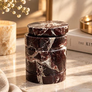 Può includere: Un contenitore cilindrico con coperchio abbinato, realizzato in marmo bordeaux scuro con venature bianche e grigie. Il contenitore presenta una finitura liscia e lucida e una distinta texture a scanalature orizzontali sul corpo esterno.
