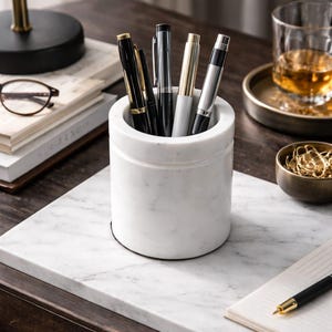 May include: A cylindrical white marble pen pot containing several pens. The pens are black, silver, and gold. The pen pot sits on a white marble surface. A notebook and a glass of amber liquid are also visible.