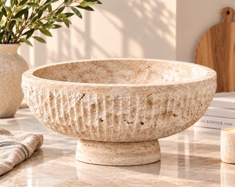 May include: A beige travertine bowl with a porous, textured surface and vertical fluted carvings along the exterior. The bowl features a wide, circular opening and rests on a sturdy, elevated pedestal base, showcasing natural stone variations.