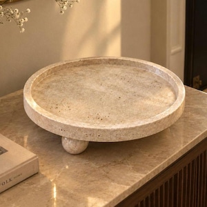 May include: A circular beige stone tray with a porous, textured surface and a raised rim. The tray is supported by three small, spherical feet made of the same light-colored, natural stone material, creating a low-profile, elevated display base.