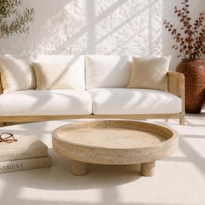 May include: A round, beige, textured stone tray with three short, cylindrical legs. The tray is positioned on a flat surface next to two stacked books titled Pride and Prejudice and Atonement, with a pair of brown-rimmed glasses resting on top.
