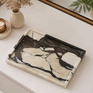 Puede incluir: Una bandeja rectangular hecha de piedra pulida con un patrón de alto contraste de vetas de color negro profundo y blanco crema. La bandeja tiene una superficie lisa y plana con bordes ligeramente elevados, mostrando la textura natural del mármol.